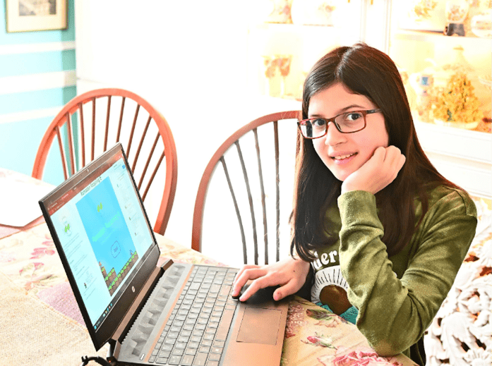 A girl sits at a table in front of a computer which has her online recycling game on the screen. 